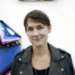 A woman with short hair in a black leather jacket, Gallerist Sadie Coles, stands confidently before vibrant artwork.