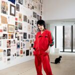 A woman, artist Sue Webster, stands in a striking red jumpsuit in a room with a black cat and art on the wall.