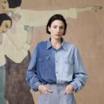 A photo of artist Sara Berman wearing a denim shirt standing in front of a large painting of two women.