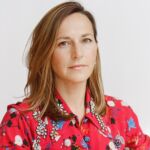 A woman dressed in a vibrant red floral shirt stands against a white backdrop, exuding a stylish and modern appearance.