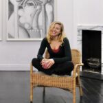A woman, artist, Gianna Dispenza, sitting cross-legged on a chair in the middle of a room in front of an artistic painting.