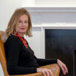 A woman with light hair, Julia Peyton-Jones, sits on a brown leather chair by a fireplace, smiling at the camera.