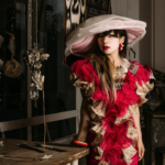 A woman leans on a wooden table wearing a dramatic red and gold dress with frills and an oversized hat.