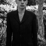 A man poses for a black and white picture in a woodland area, wearing a black suit and cross necklace.