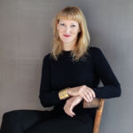 A woman with long blonde hair, curator Emma Ridgway sits with her hands crossed on a wooden chair, wearing a black outfit smiling at the camera.