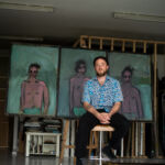 A man, painter Jack Penny, sits on a stool in front of three paintings of men wearing sunglasses.