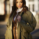 A woman with dark hair, Katy Wickremesinghe, stands outside with the sun on her face wearing a green jacket, brown and white turtleneck.