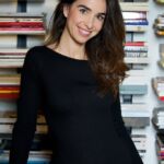 A woman wearing a black top and jeans stands confidently in front of a large bookshelf filled with various books.