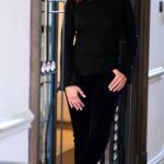 A woman, art collector Maria Sukkar, stands poised in black trousers and a turtleneck sweater, positioned in front of a door.