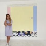 A woman with blonde hair, artist Fleur Yearsley, stands in front of a large painting of a colourful bathroom cubicle, wearing a lilac dress.