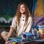 A woman with long, red, curly hair, artist Atalanta Xanthe, sits in a white dress smiling at the camera with paint scattered across the table and colourful artwork behind her.