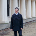 A woman with short grey hair, Bengi Ünsal, stands outside in front of a white building, wearing a navy jacket and trousers.