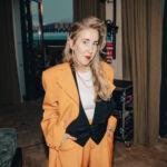A woman in a striking orange suit, Gemma Rolls-Bentley, chief curator at Avant Arte, confidently poses for the camera.