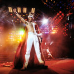 A man, Freddie Mercury, stands on stage with bright lights behind him wearing a crown and a grand shawl with his arm in the air.