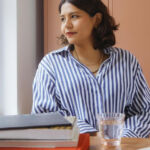 Tarini Malik, an art curator, is seated at a table with books and a glass of water, wearing a striped shirt, looking wistfully out the window.