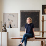A woman with blonde hair, Artist Teniqua Crawford, sits on a table with her legs crossed surrounded by paintings and sculptures.
