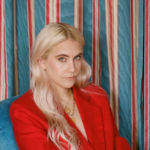 A blonde woman, India Rose James, sits on a blue chair wearing a vivid red jacket in front of rainbow background.