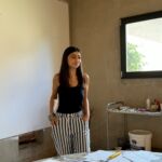 A dark haired woman, visual artist Irini Bachlitzanaki, stands in an art studio with her hands in her pocket wearing black and white stripey trousers and a black top.