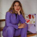 Portrait of blonde woman, artist Leila Lallali, sitting on a staircase wearing a purple jumpsuit