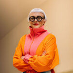 Woman with a silver bob hair cut, designer Morag Myerscough, wearing black glasses and a colourful bright orange and pink jacket