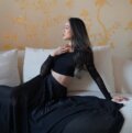 Brunette asian woman, architect Dara Huang, in all black outfit looking to the right – seated on a white sofa in front of a yellow floral wallpaper