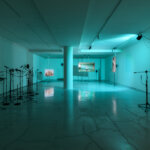 Gallery exhibition, Claridge's ArtSpace, lit in light blue with three multi-channel videos in the distance, and a group of speakers