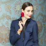 Brunette woman, fashion entrepreneur Maria Kastani, smiling in red lipstick and red circular earrings against a light floral blue wall.