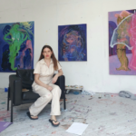 Brunette woman in beige jumpsuit, artist Kate Lyddon, sitting on black chair in front of colourful paintings.