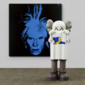 KAWS figure holding a white tea shirt with a Warhol banana and blue blue cartoon lying down behind, with a black and blue Warhol self portrait behind.