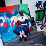 A man, artist Slawn, sits on a chair in an art studio amongst various bright graffiti paintings wearing a white t-shirt, blue shorts, a green cap, and leopard print loafers.