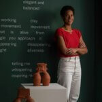 A woman with short dark hair, Bisila Noha, stands against a green wall with white writing on it wearing white jeans and a red top, with her ceramic pots on display.