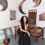 A woman with dark hair, Charlotte Edey, wears a black jumpsuit and sits on a wooden chair in an art studio surrounded by various pieces of art.
