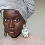 A close up of a woman's face, Dola posh, wearing large silver and gold earrings, a soft grey head wrap and pink roll neck top.