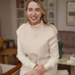 Blonde woman, Jenn Ellis, smiling in full beige outfit sitting on mid-century modern chair