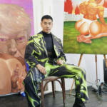 A man, John Hui, confidently sits on a chair with abstract paintings behind him, wearing green and black patterned suit and doc martens.