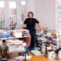 A woman, Sutapa Biswas, stands in a room amongst a lot of books wearing black t-shirt, navy trousers, and blue crocs.