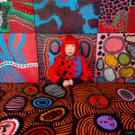 A woman with red hair sits in amongst vivid, multicoloured paintings.