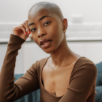 Bald black woman, Africa Brooke, with red nail polish and wearing a brown long sleeve shirt looking into the camera