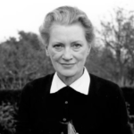 A black and white photo of Elizabeth 'Lee' Miller, wearing a blacking cardigan and white collared shirt standing in front of a hedge and some plants.