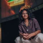 A woman with dark hair, Meneesha Kellay, sits with her hands clasped wearing a brown shirt and light trousers with a large screen projected behind her.