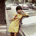 Vintage photograph of a Black woman wearing a psychedelic short dress with short hair, resting against a grey car