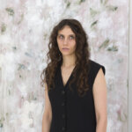 Portrait of a brunette woman, Bianca Raffaella, in a sleeveless blazer, in front of an abstract painting
