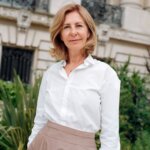 A woman, Florence Bourgeois, stands outside against some green plants wearing a peach skirt and white shirt.