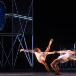 Two dancers in tight white outfits holding each other's hands and extending outwards