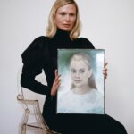 Blonde woman in black dress, Hilde Lynn Helphenstein, sitting on a plastic chair holding a painting of a young girl