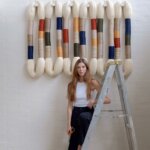 Blonde woman in white tank top, Alejandra Aristizabal, leaning against a ladder in front of a woven textile artwork