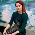 White woman with short red hair, Laura Footes, sitting with smock in front of abstract painting