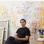 Portrait of man in all black, Rob Davis, sitting in front of a painted colourful wall