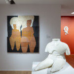 Exhibition photograph of a white nude female body without a head at the forefront, and a painting of two figurative women behind
