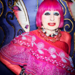 Woman, Zandra Rhodes, with pink hair, colourful eye makeup and red and pink outfit looking at the camera